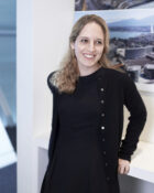 A photograph of Mina Hasman, a smiling woman with light brown hair and wearing a black dress and cardigan.