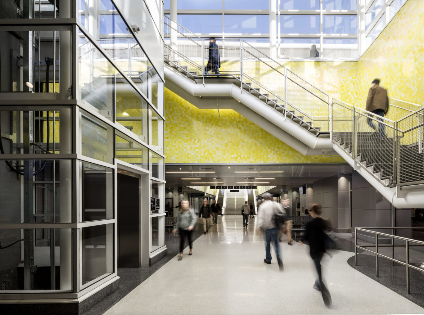 Slide 2 of 7, Underground, a 22-gate bus concourse services 16 regional, express, and local bus routes. The terminal, measuring 980 feet in length, serves a dual purpose as a pedestrian concourse that connects the constellation of transportation programs distributed across the site. Vivid colors and natural lighting help passengers orient themselves while handsome terrazzo floors and sparkling yellow glass tilework elevate the ambience of terminal beyond the often-unimaginative bus depot experience. A series of skylights and glass pavilions flood the hall with daylight, infusing the station with a sense of motion and spaciousness. One of the largest projects of its kind in the United States, the redevelopment of the former rail yards at Denver Union Station is a case study of the power of transit-oriented urban design. This substantial public investment has catalyzed an unprecedented wave of private-sector activity. Sensitive to its historic location, but fundamentally forward looking in its technical sophistication and city-building spirit, Denver Union Station sets the standard for 21st-century intermodal hubs.